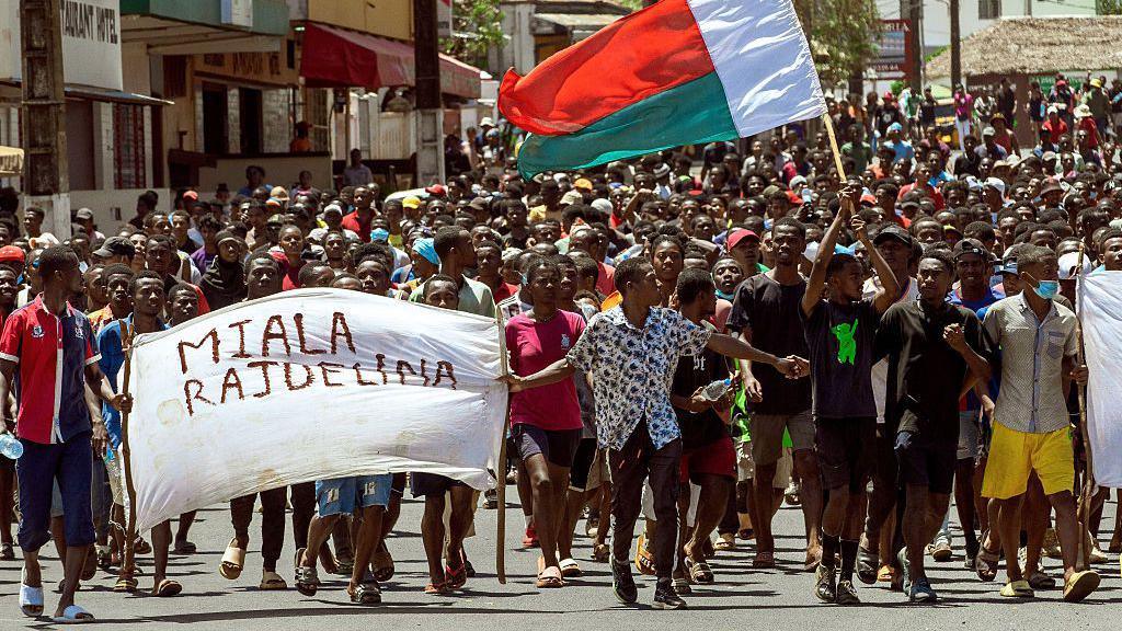 Dozens of protesters can be seen marching down a road. At the front are people holding a banner which says 'Rajoelina resign' in Malagasy. A Madagascan flag can also be seen. 