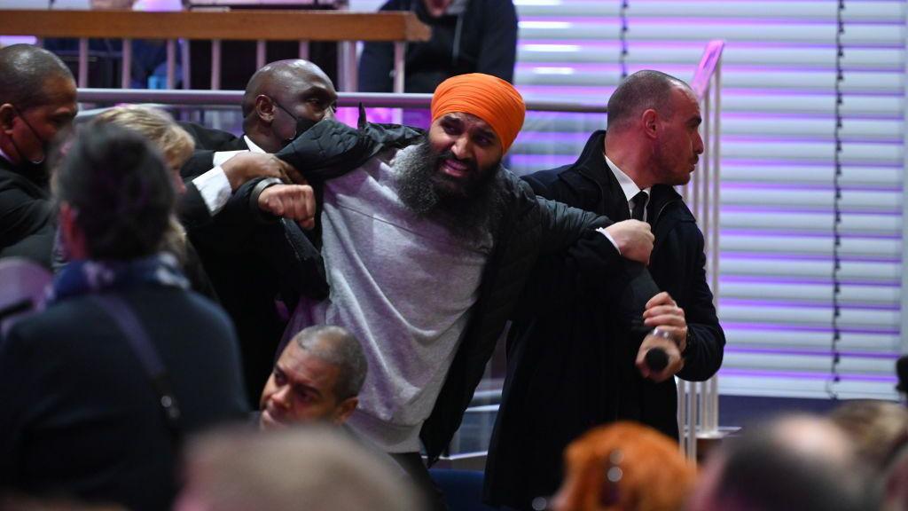 An angry man in a grey sweatshirt, black jacket and orange turban is being dragged out of a public meeting by two security staff dressed in black. They are holding his arms back as the protesting man shouts. 