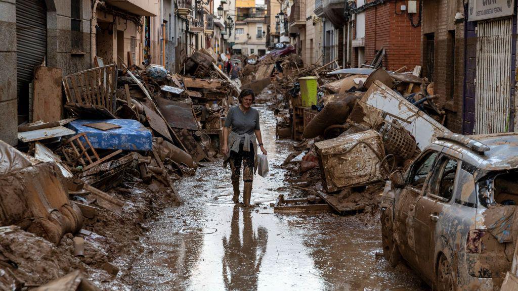 A woman walking down a flooded street. There's piles of debris including furniture and cars on both sides of the street, with buildings lining each side.