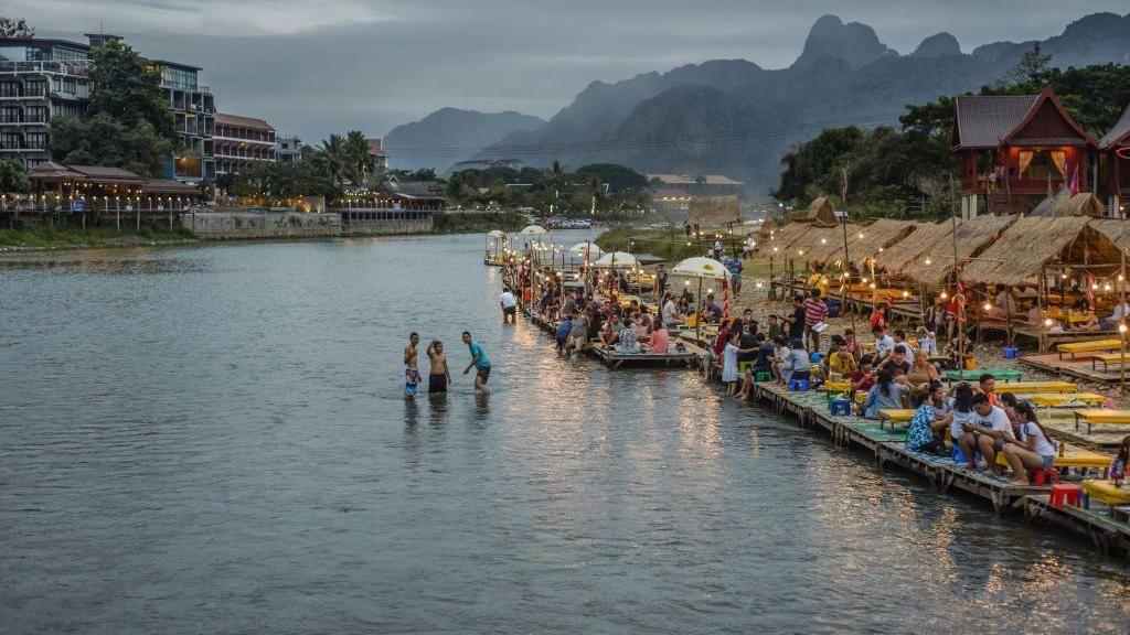 Wide view of a river in Vang Vieng. Three people can be seen in the river while crowds are seen sitting along the river front. Mountains can be seen in the background with the town's skyline