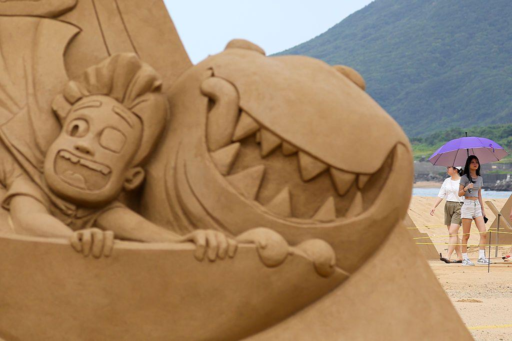 A lady with an umbrella standing behind a sand art pyramid of Elio characters