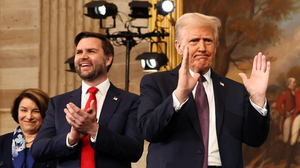 JD Vance in blue suit and white shirt with red tie standing smiling and applauding while, to his right, Donald Trump is gesticulating to somebody off camera. The president is wearing a dark blue suit, white shirt and deep burgundy patterned tie. They are in a neo-classical building.