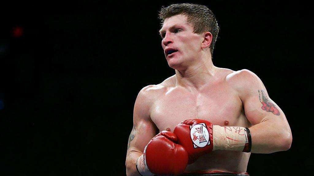 A tattooed man is mid-fight and shirtless, with red boxing gloves tapes onto his hands. 