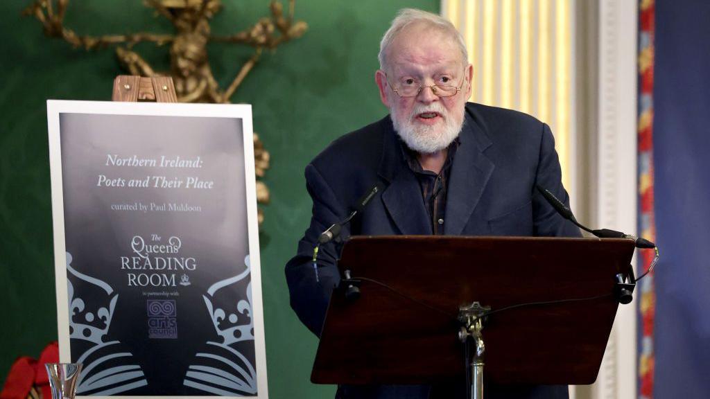 Michael Longley, with short white hair and beard, wearing glasses, wears a dark suit and stands behind a lectern. To his right is a sign saying "Northern Ireland Poets and Their Place".