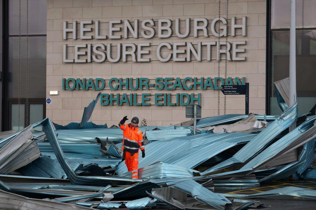 A man in orange overalls is walking on top of the metal roof of the leisure centre and the roof is in the car park, badly damaged.