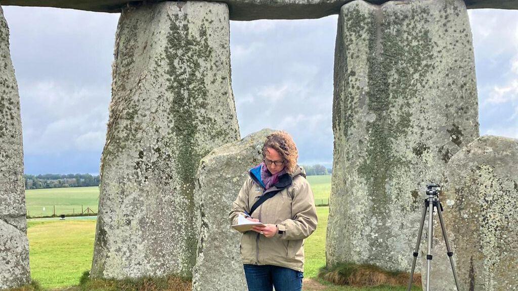 Rose Ferraby jotting down some notes while studying Stonehenge. She is standing in front of several of the standing stones, but is within the circle, rather than outside it. The clouds are a moody grey and the sunlight is breaking through onto the ground.