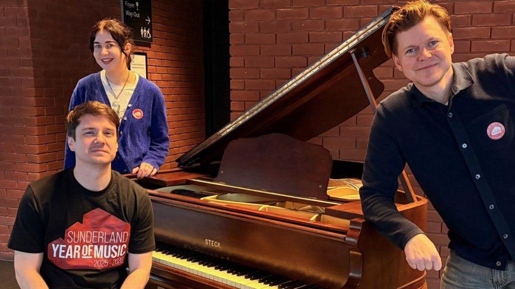 Three representatives from Sunderland Music City gathered around a brown grand piano which will be used during the concert.