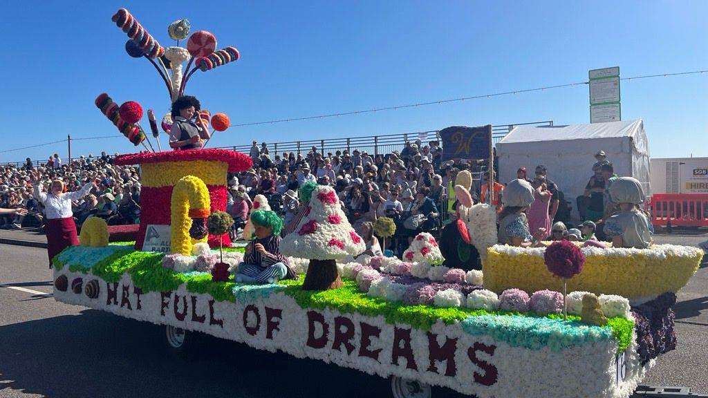 A flower display at the Battle of the Flowers event in 2024. The display says on the side 'Hat full of dreams' and there is a red and yellow hat on the left, mushrooms and candy canes made out of flowers.