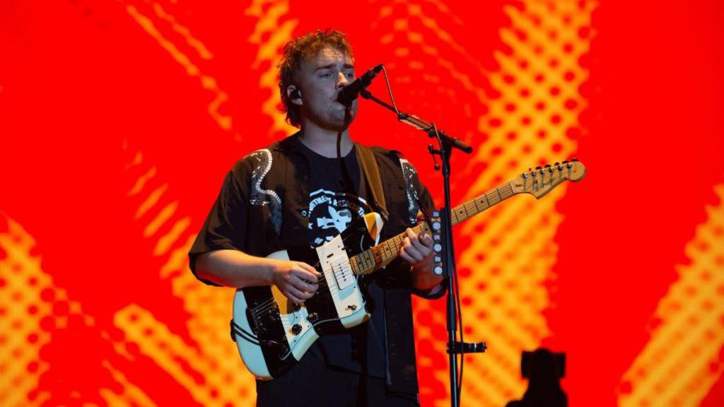 A face on photo of a singer holding a guitar. The background is red from a projector. He is wearing a black top and has curly short hair.