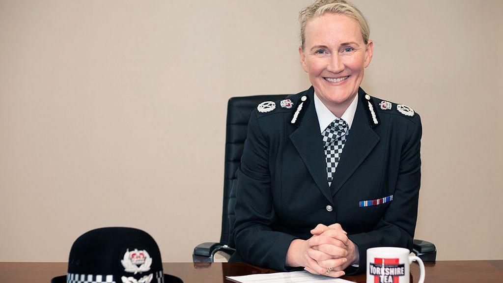 A smiling Serena Kennedy, chief constable of Merseyside Police, who has blonde hair, sits at a brown wooden desk in her black uniform with her police hat and a mug branded with Yorkshire Tea in front of her. 