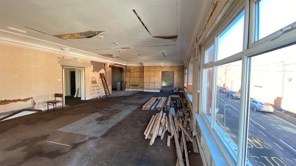 Interior of a room which has been stripped out. There is exposed plasterwork on the walls two visible walls, a damaged ceiling, and stacks of wood on the floor. to the right is a large window overlooking a road and some parked vehicles