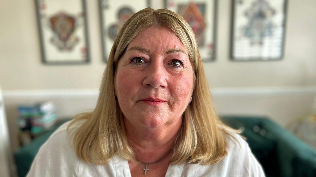 Portrait photo of Rachel Osmond, looking directly at the camera, with a slight smile on her face. She has straight, shoulder-length hair with blonde highlights. She is wearing a white blouse and a crucifix necklace. She is pictured in a living room, with a sofa and framed wall pictures in the background. 