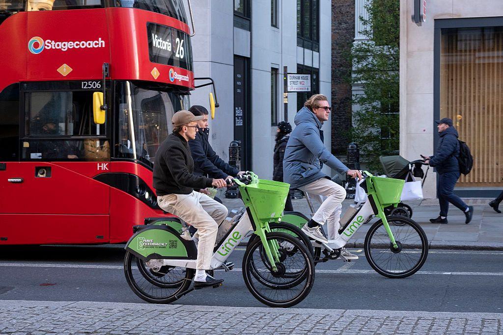 Two Lime bikers ride in front of a London bus on a city centre street.