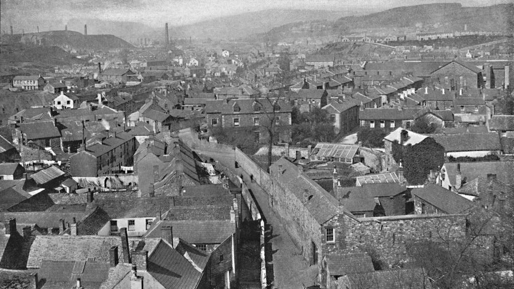 Merthyr Tydfil, circa 1896, it is a view of the town in black and white with densely packed terraced housing and industrial chimneys in the distance. Hills can be seen in the distance.