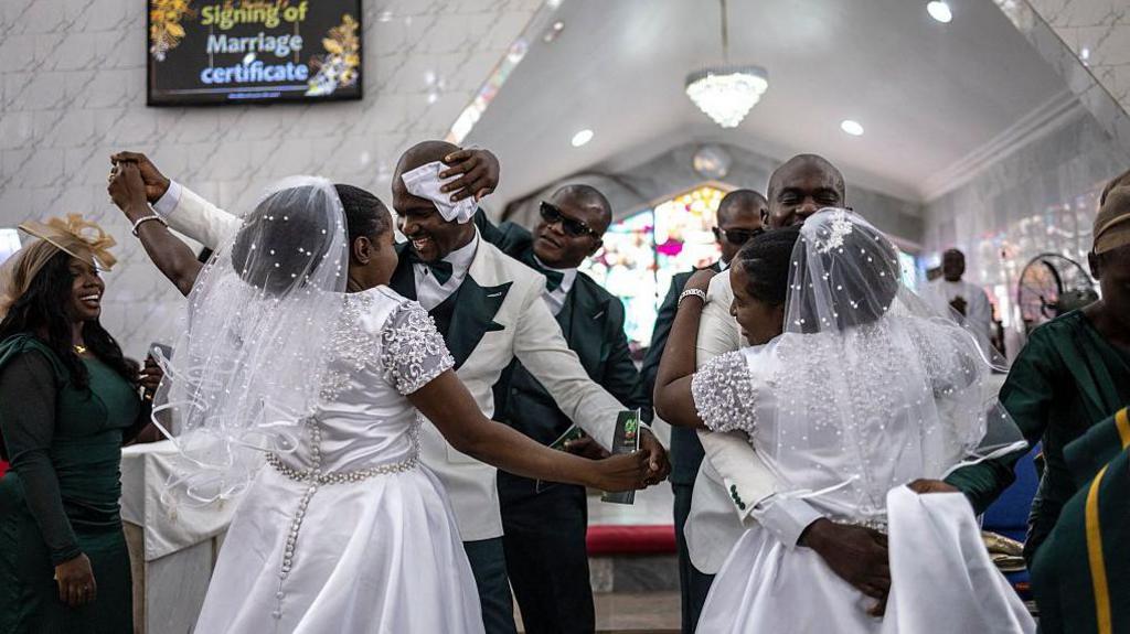 The grooms and brides dance during their wedding ceremony. A man wearing sunglasses wipes one of the groom's brow with a white cloth.