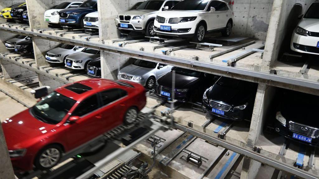 A high-tech, multi-level automated car park with concrete walls and tracks. Multiple cars are neatly slotted into mechanical platforms, while a red sedan is being transported on a motorised rail system in the foreground.