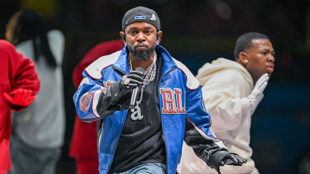 Rapper Kendrick Lamar wears a blue varsity jacket and backwards baseball cap while performing in front of dancers at the 2025 Super Bowl half-time show.