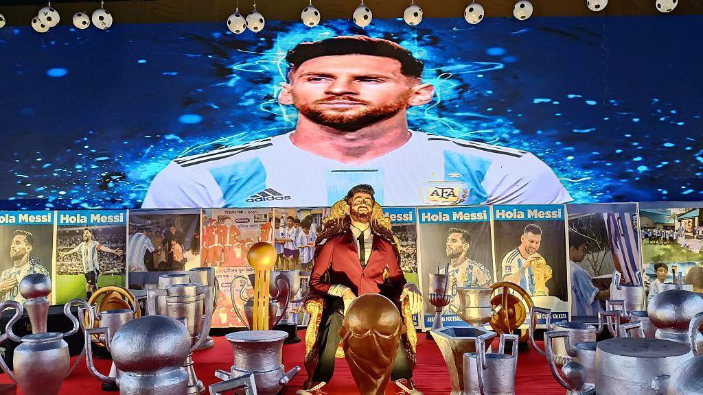 A life-sized Lionel Messi sits on a throne next to replica trophies and a large image of Lionel Messi on the wall