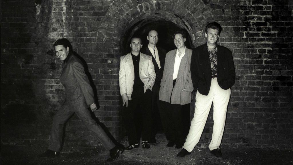 A black and white image of the band Squeeze, the five male members of the band wear oversized suits and are posing in what looks like an underground tunnel with an archway behind them.