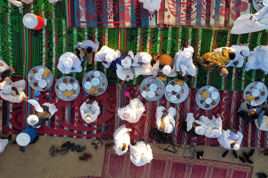 An aerial image of several Sudanese men sitting on colourful rugs as they prepare to eat the food spread out on multiple platters - Friday 7 March 2025.