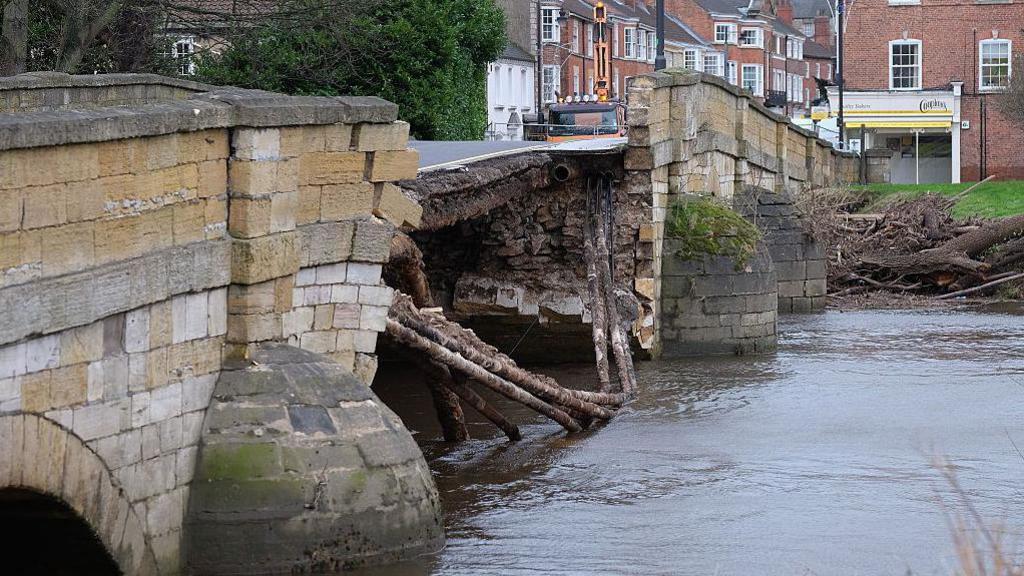 An old stone bridge with one side of it in the water of the river below after a partial collapse.