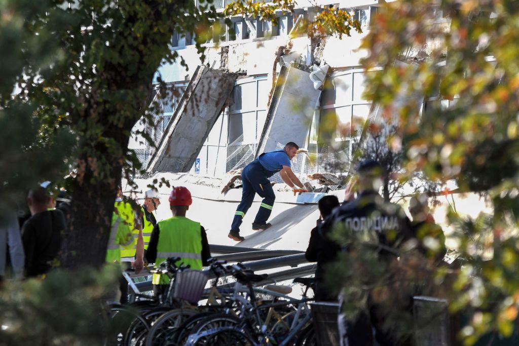 A rescuer leans into and appears to pull on part of concrete that collapsed outside a railway station in Serbia while rescuers in hard hats and police officers look on 