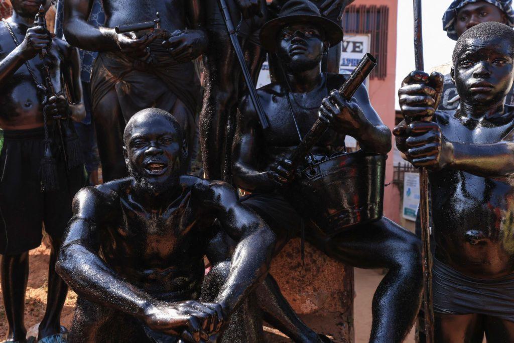 Topless men painted in black body paint crouch together. Their expressions are stern and some carry ceremonial weapons in Foumban, Cameroon - Sunday 8 December 2024