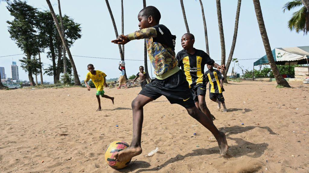 Barefoot boys in football kits play football on a sandy pitch in Abidjan. Palm trees can be seen in the background - Wednesday 26 February 2025.