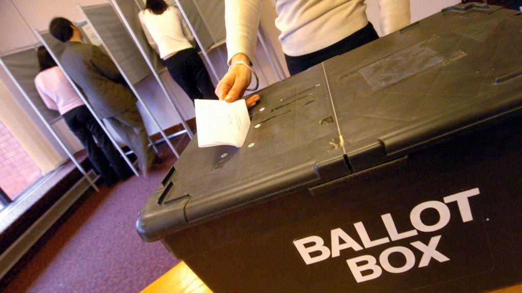 A black ballot box on a table with someone putting their ballot paper through the slot on top