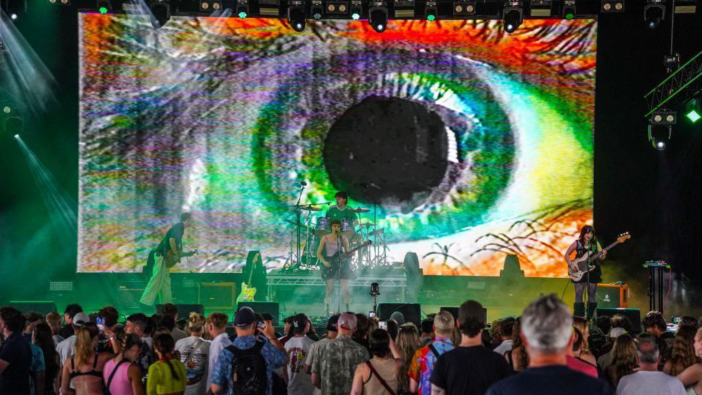 A band performs to a crowd on a stage, the screen behind them shows a colourful eye