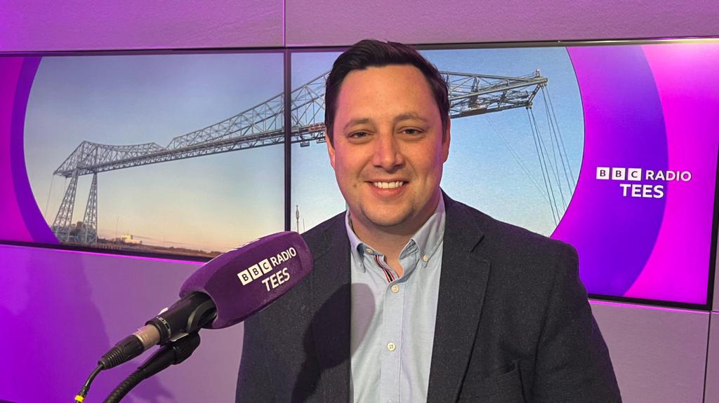 Ben Houchen sitting in the studio at BBC Tees. He is wearing a light blue shirt and a dark jacket, has short dark hair and is smiling. In front of him is a microphone with a purple cover and the BBC Tees branding in white letters. Behind him is a wide screen with the BBC Tees logo in white lettering on a purple background and an image of the Transporter Bridge