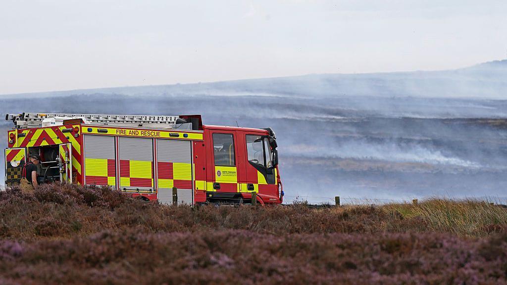 A fire engine parked on Langdale Moor. Heather can be seen in the foreground, with smoke in the distance.
