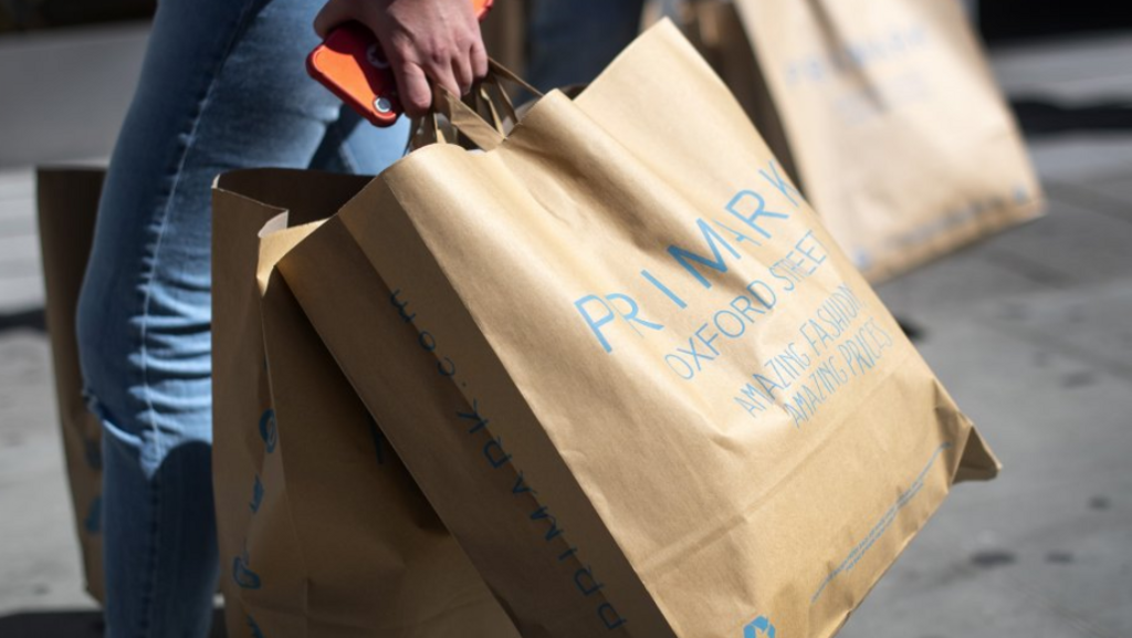 A hand is seen holding two brown Primark bags. The pavement is visible in the background.