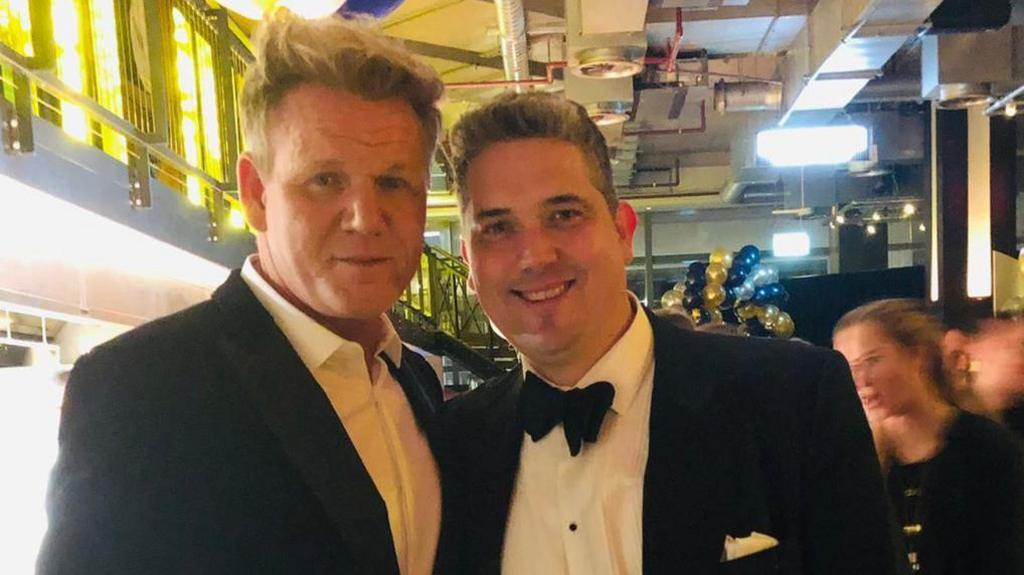 Two men looking at the camera and smiling. The man on the right is wearing a black suit and a black bowtie, and he has short brown and grey hair. The man on the left is also wearing a black suit jacket and has short blond hair.