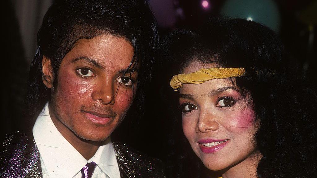 Michael Jackson in a white shirt, dark sparkly tie and dark sparkly jacket, next to La Toya Jackson who is wearing a yellow headband. 