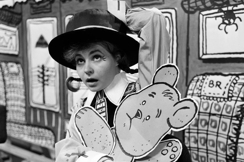 Prunella Scales photographed in one of her earliest acting roles in 1962. She is dressed as a school girl with pigtails and a boater hat.  She is carrying a cut-out of a teddy bear.