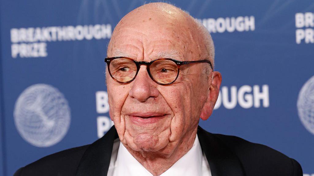 An elderly Rupert is pictured wearing glasses and a black suit jacket.