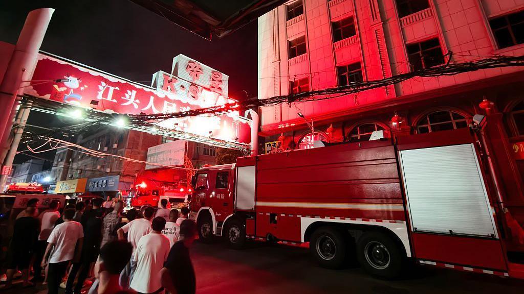 A crowd stands beside a red firetruck in the south-eastern Chinese city of Jinjiang where a massive fire killed at least 28 people