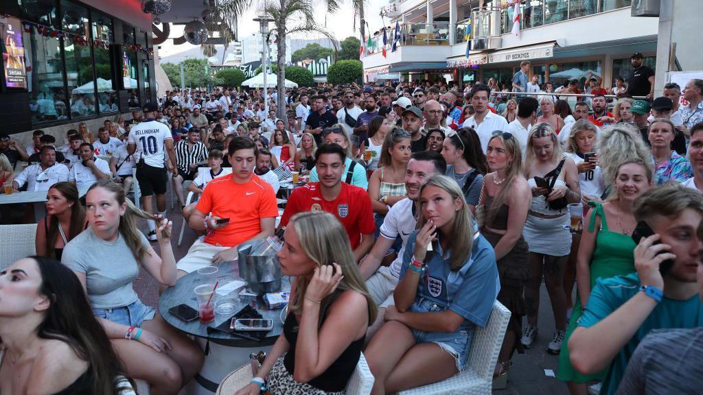 People sit at tables or stand in a crowded street with buildings on either side. Many are looking up towards a screen and some are checking their phones