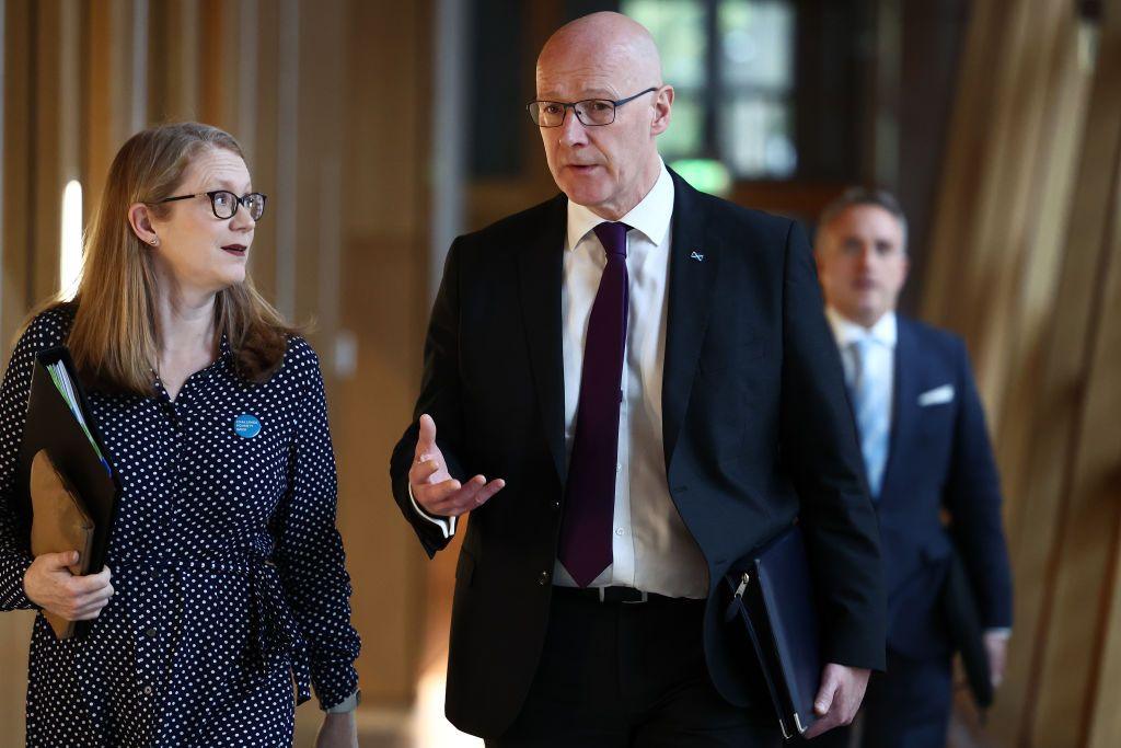 Shirley-Anne Somerville (left) is walking, carrying a black document folder, beside John Swinney in a corridor at Holyrood. Alex Cole-Hamilton can be seen in the background over Swinney's shoulder.