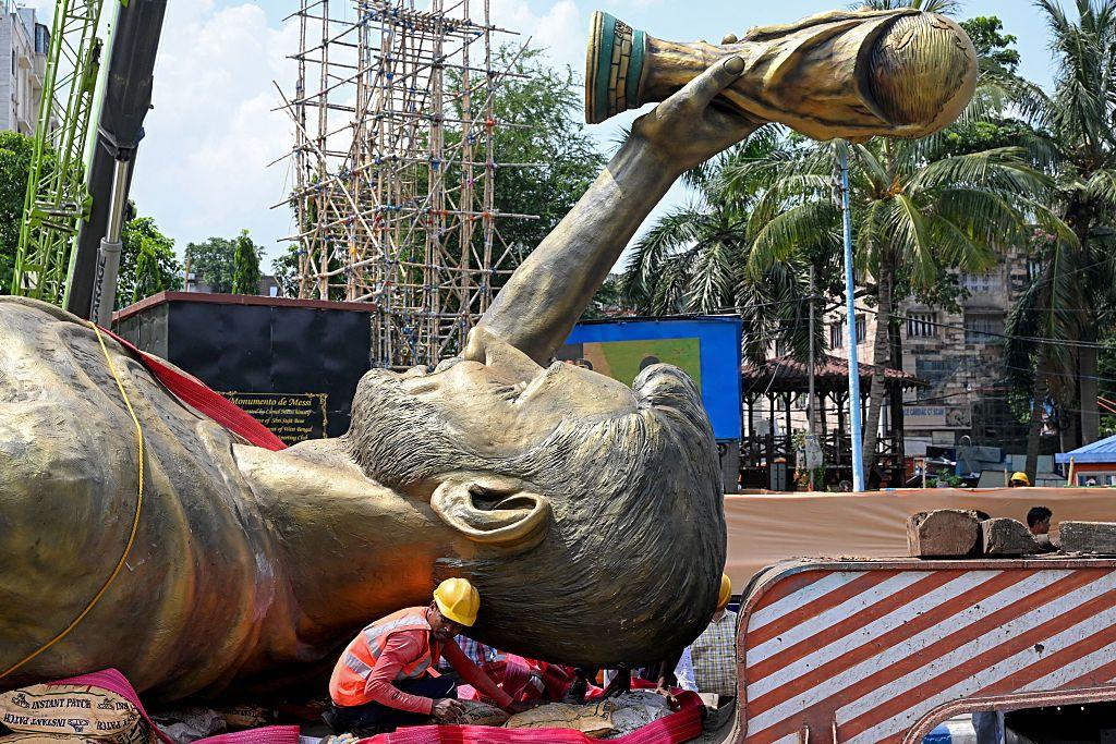 Lionel Messi's statue lays flat as one builder works on securing it. You can see the World Cup aloft above the statue's head.