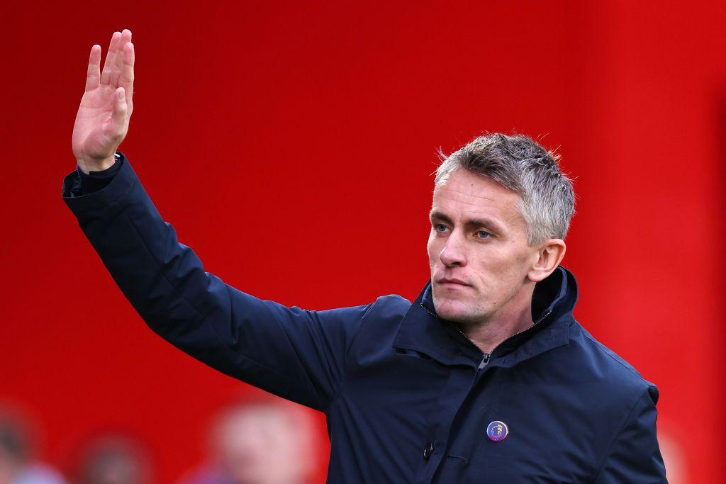 Ipswich Town manager Kieran McKenna raising his right hand in a wave or gesture, wearing a dark blue jacket with a Premier League badge, set against a bright red stadium background.