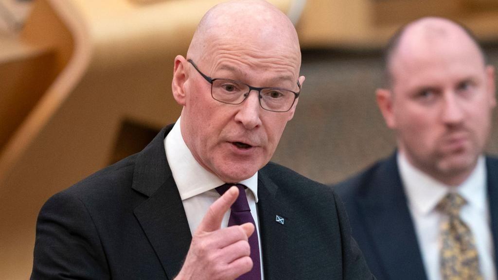 First Minister John Swinney gesticulates with his left index finger with Health Secretary Neil Gray sitting in the background
