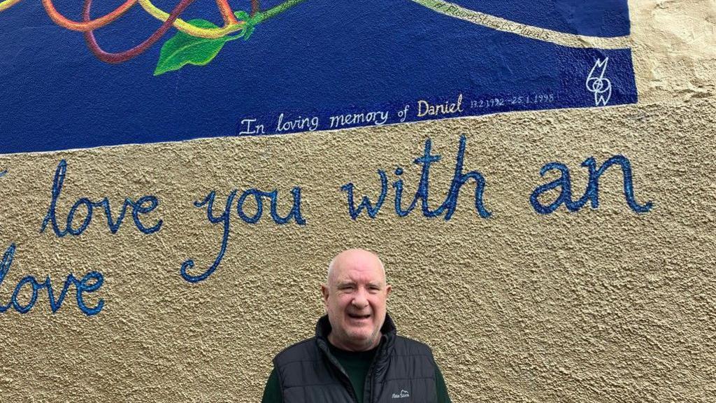 David Brown is smiling, wearing a black gilet jacket and green top, as he stands in front of the mural which pays tribute to his late son. The words 'In loving memory of Daniel 17.2.1992 to 25.1.1998' can be seen.