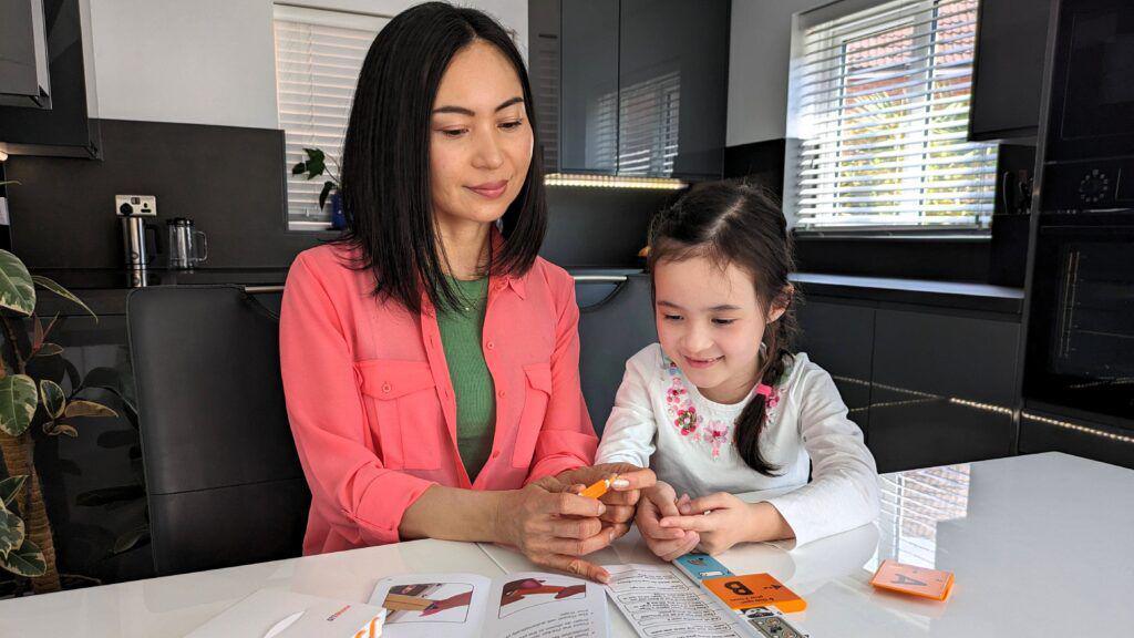 A woman and a child are using looking at the test kit while sat at a table in a black and white office space. The woman is wearing a coral shirt with a green top underneath, while the child wears a white long-sleeve top with pink flowers around the neckline.