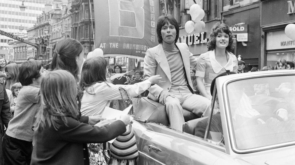 Cliff Richard and Deborah Watling in an open top car with crowds of people asking for autographs