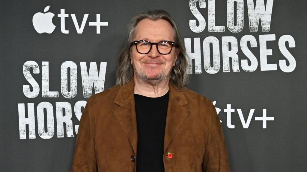 Gary Oldman is wearing a brown jacket and black jumper underneath. He has long mostly grey hair and is unshaven resembling his Slow Horses character Jackson Lamb. He is at an event promoting Slow Horses and standing next to the show's logo.