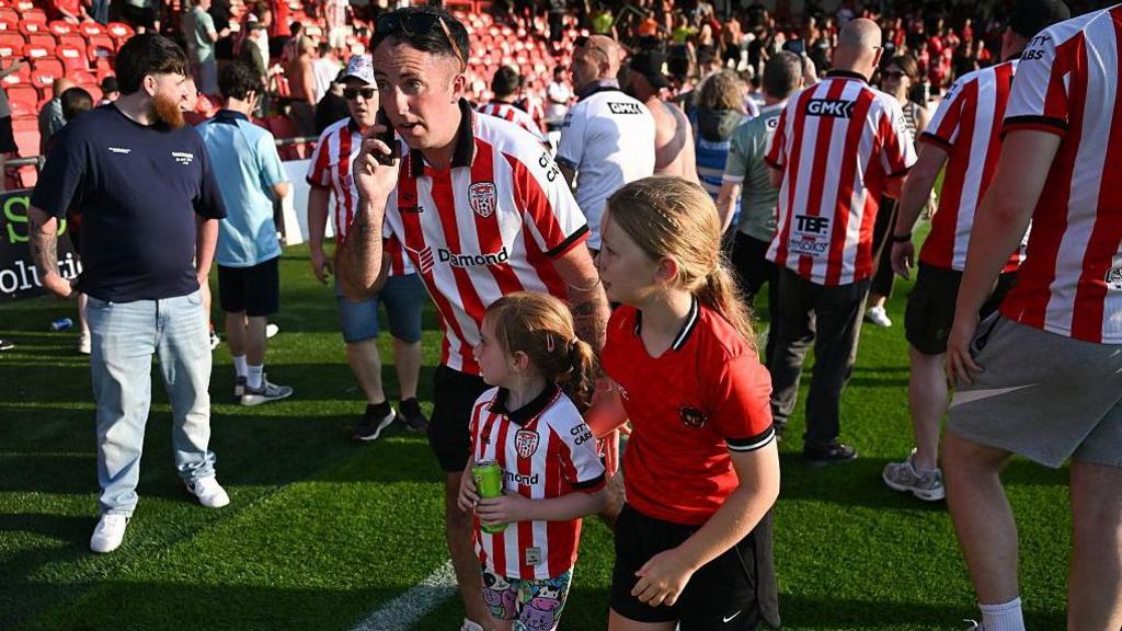 A Derry City supporter speaks on the phone beside two children on the pitch at the Ryan McBride Brandywell Stadium