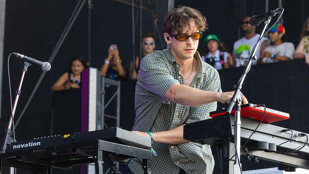 Barry Can’t Swim performing on stage, wearing sunglasses and a checked shirt, playing multiple keyboards and electronic equipment, with festival-goers watching in the background.