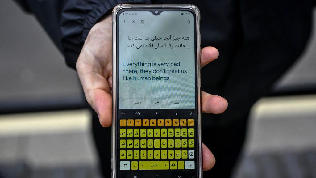 A hand holding a phone that has translated a message into English. It reads: "Everything is very bad there, they don't treat us like human beings"